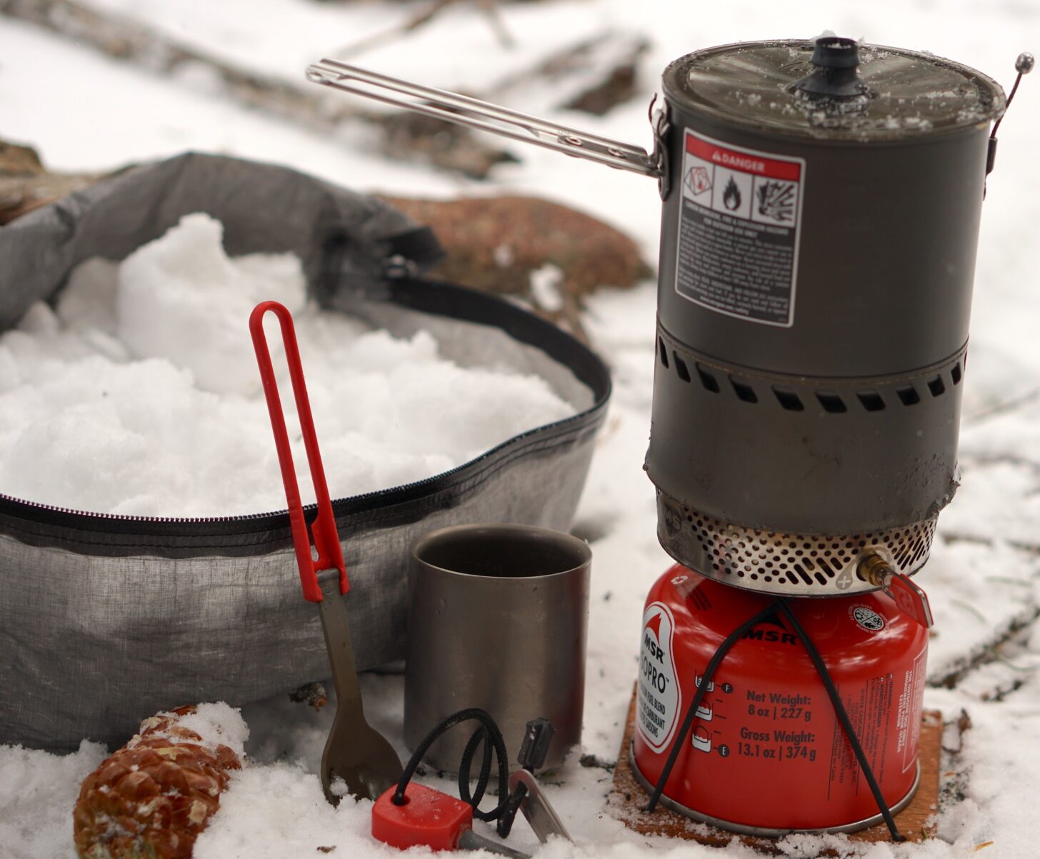 a stuff sack full of snow (to be used for melting to create water while cooking), a fuel canister, a canister stove, a titanium mug, a folding spoon, and a fire starting device
