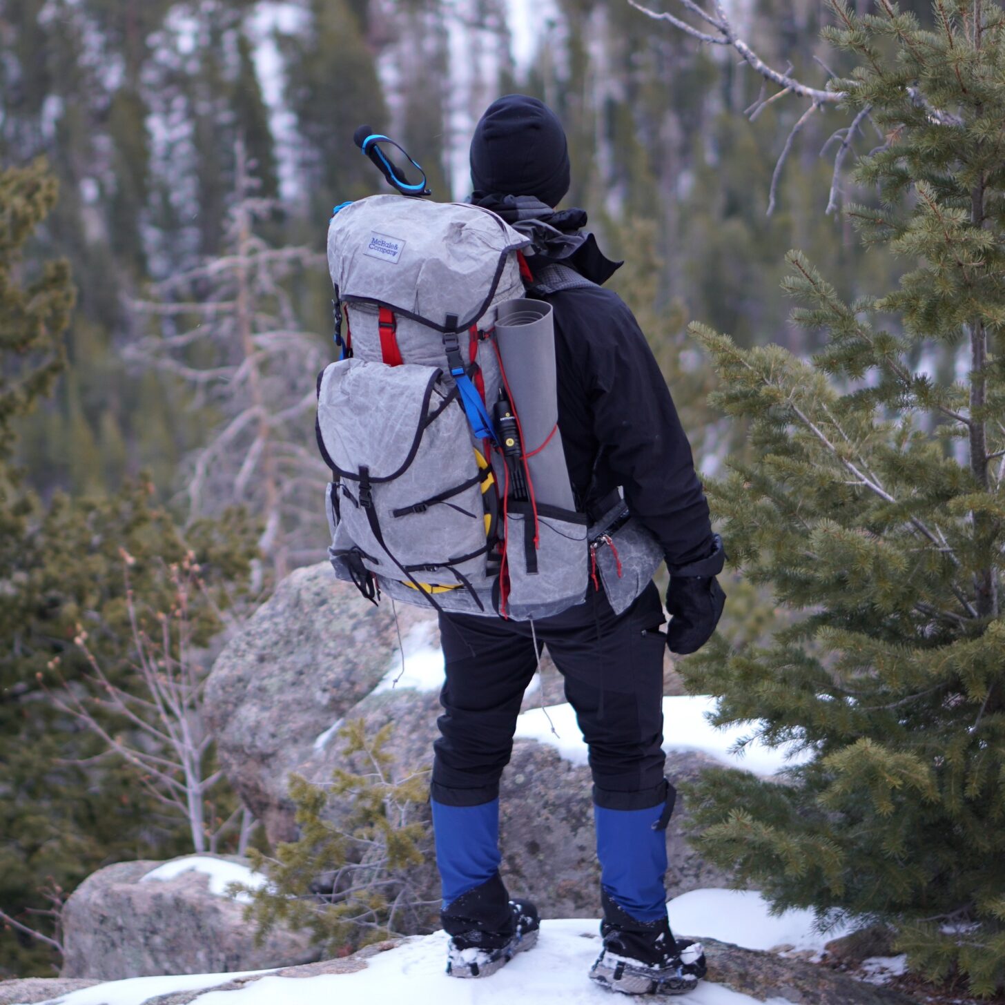 man standing, wearing a backpack on a snowy hill in the forest during the winter
