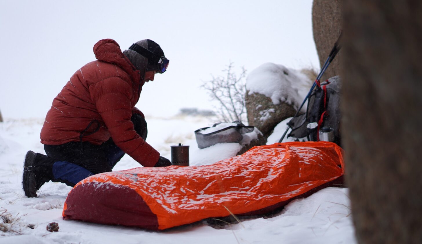 man operating a backpacking stove near his bivy sack in a snowy winter camp