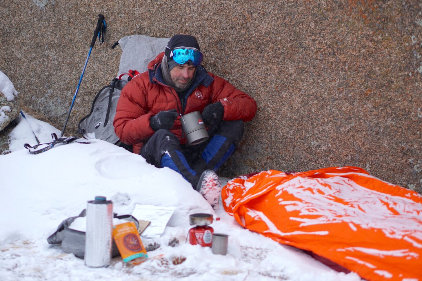 man eating soup wearing a down parka at a bivy camp in the snow