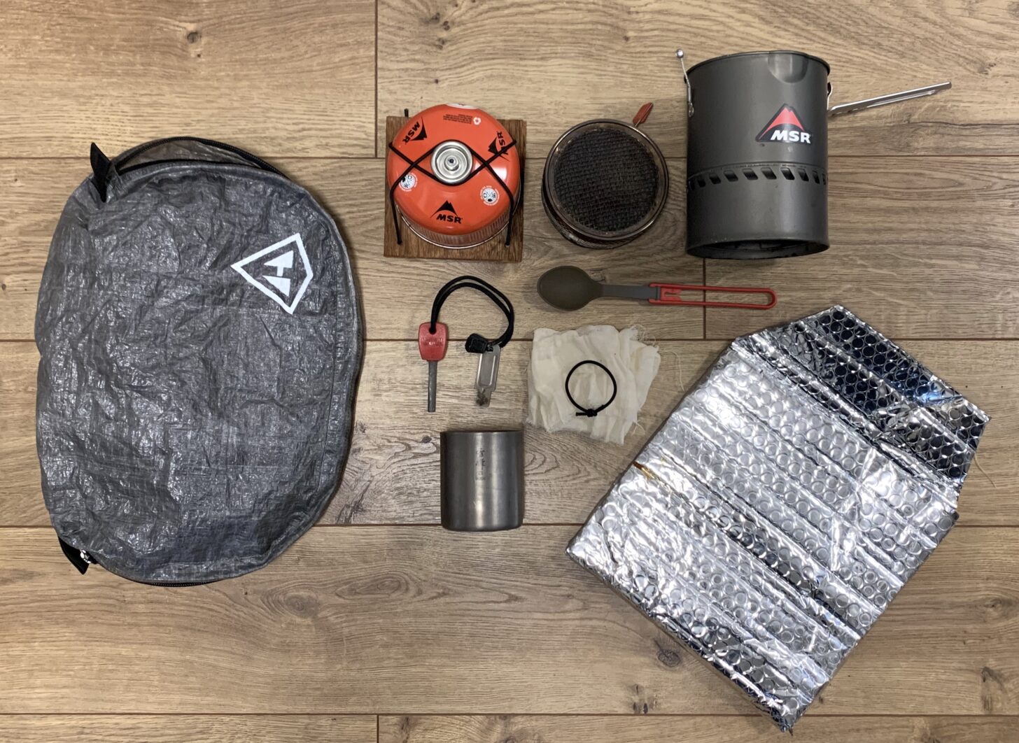 layout of cooking gear, including a zippered stow bag, fuel canister on a plywood base, radiant burner stove, heat exchanger cook pot, folding spoon, fire starter, titanium mug, cheesecloth for filtering melted snow into a water bottle, and homemade reflective bubble wrap food cozy