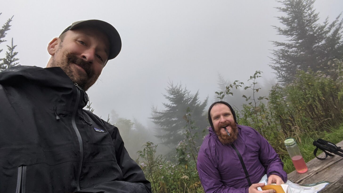 two men smiling and sitting in a backcountry location, one is taking notes in a notebook