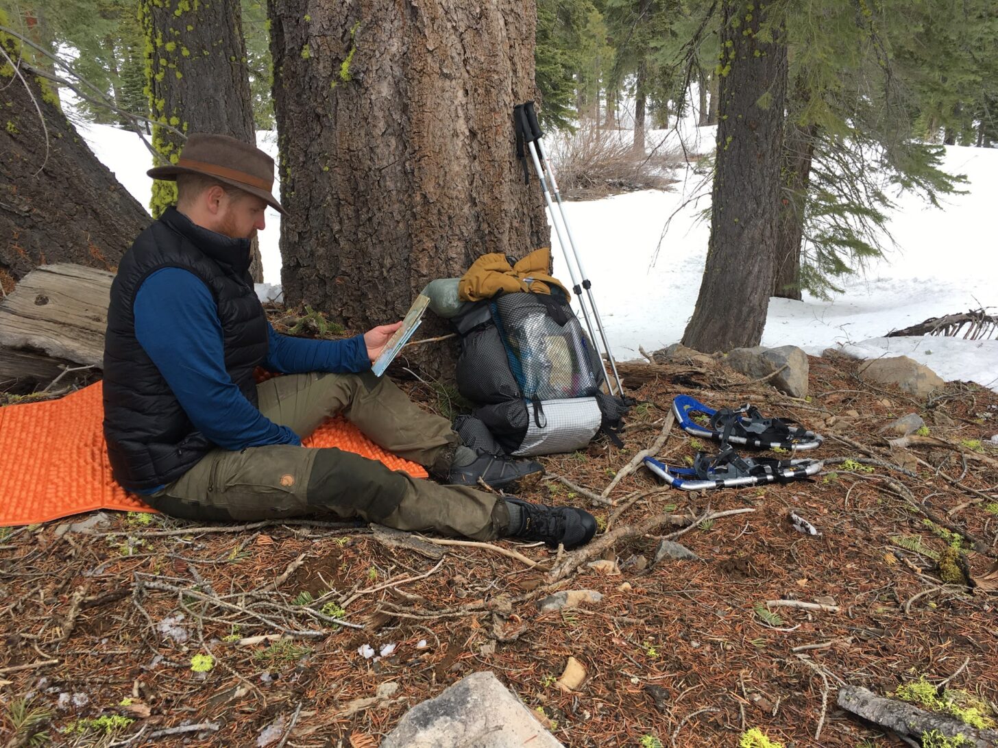 man reading a map while sitting on a foam sleeping mat, with backpack, trekking poles, and snowshoes nearby