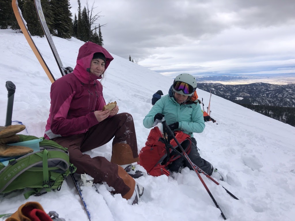 a woman eats a sandwich while sitting in snow on the side of a mountain