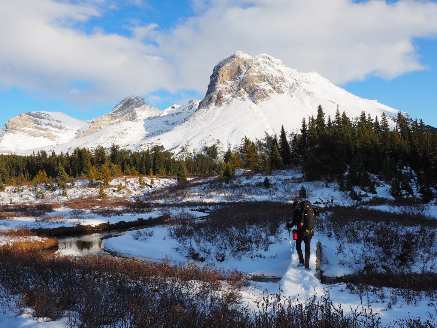 A woman crosses a snowy creek with a mountain in the background.
