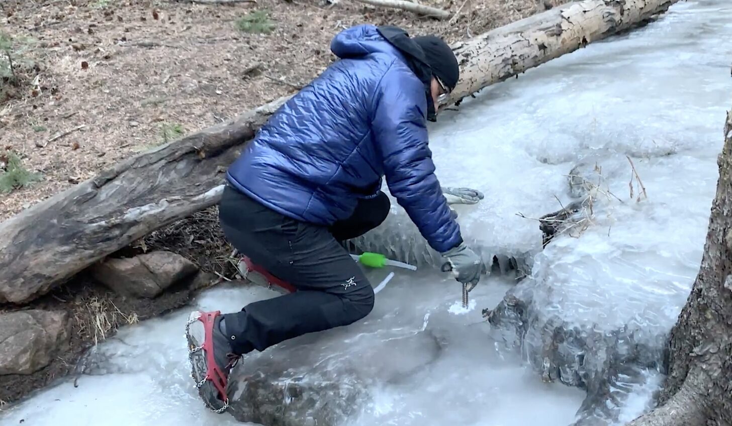 man using an ice screw to drill through a frozen stream surface
