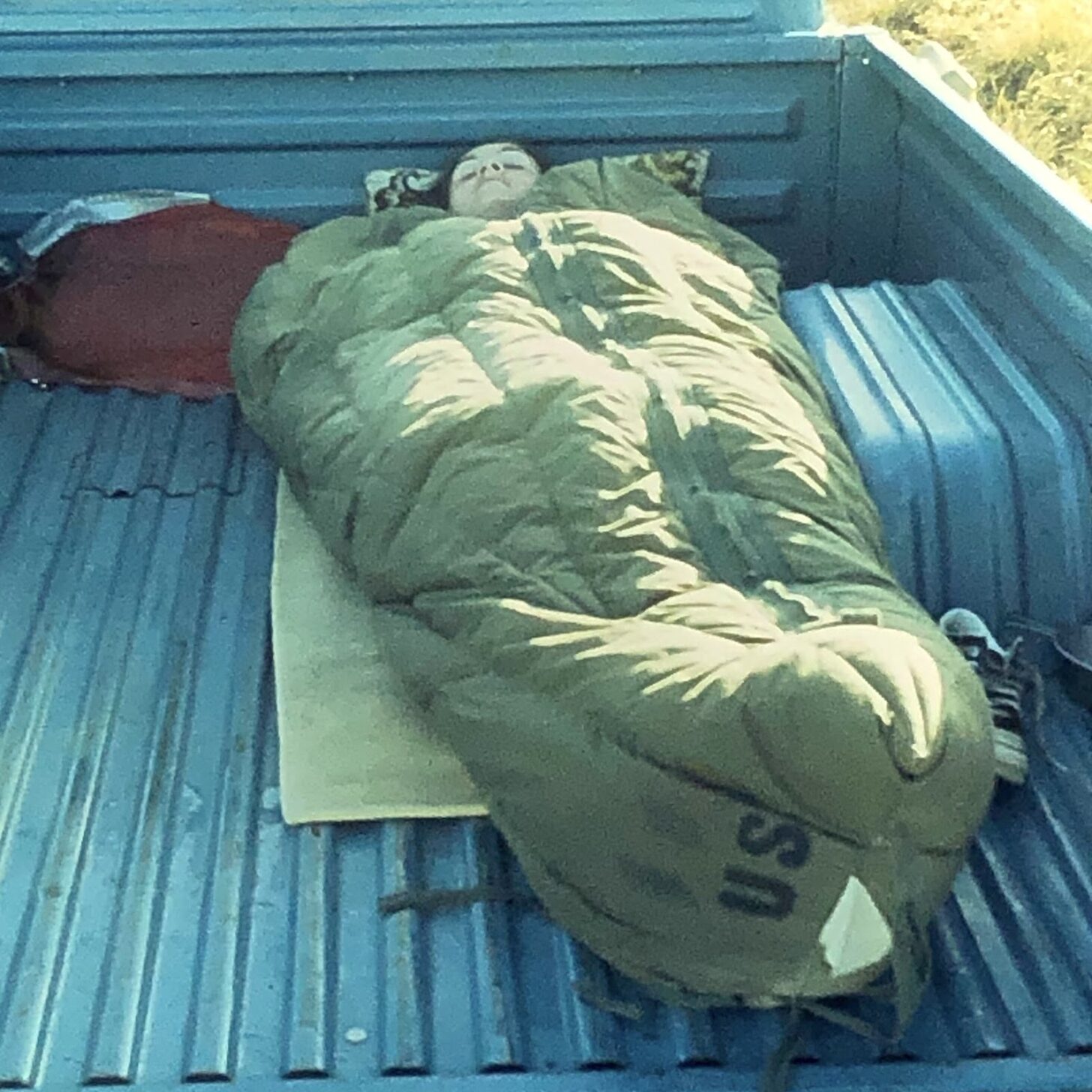 a person sleeping in an old army surplus sleeping bag in the back of a truck.