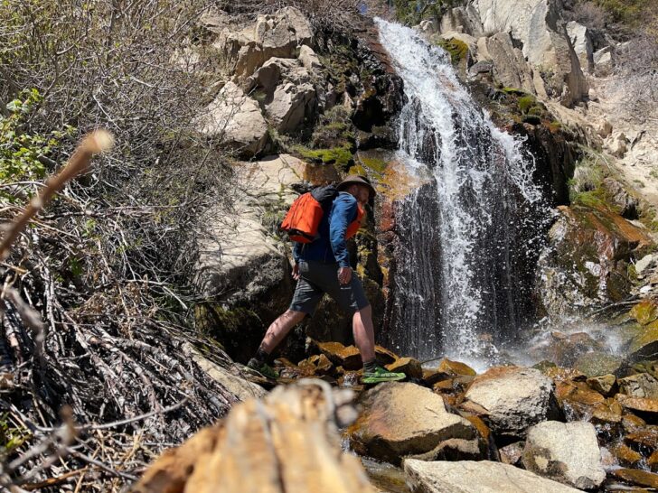 a man works his way underneath a waterfall while wearing an orange backpack.