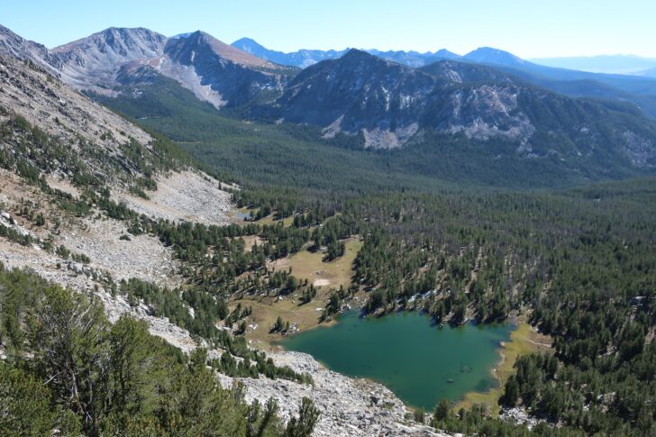 A wide landscape photo with mountains in the background. In the foreground, down in a valley, a blue lake sits like a gem in a green forest.