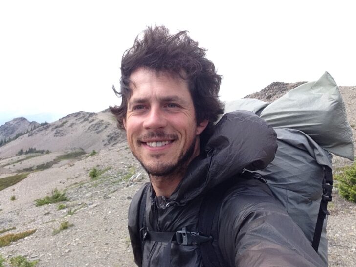 A man wearing a backpack smiles into the camera with wilderness behind him