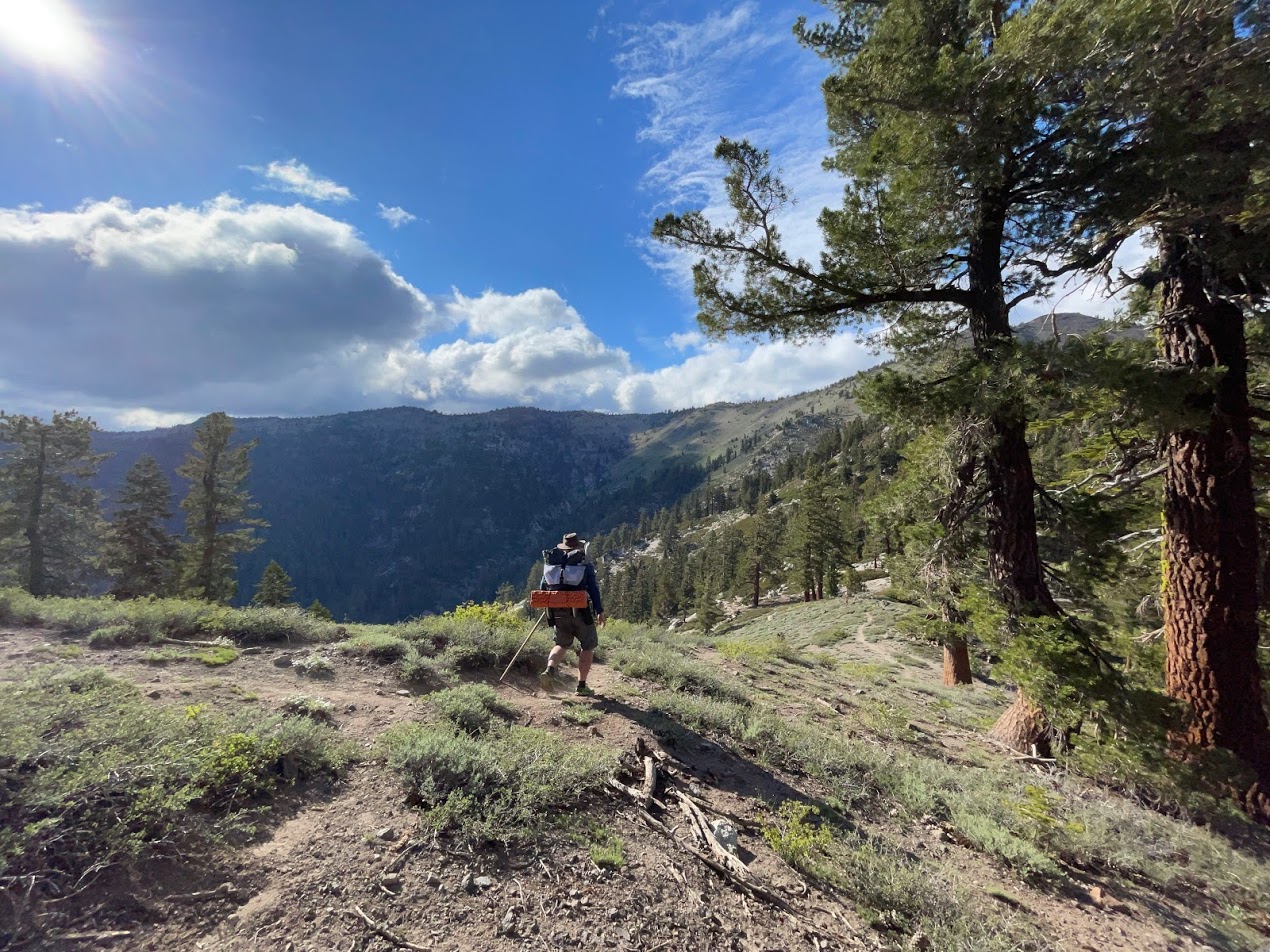 a man hikes with the pack on a ridge with a mountain view and a valley in the background.