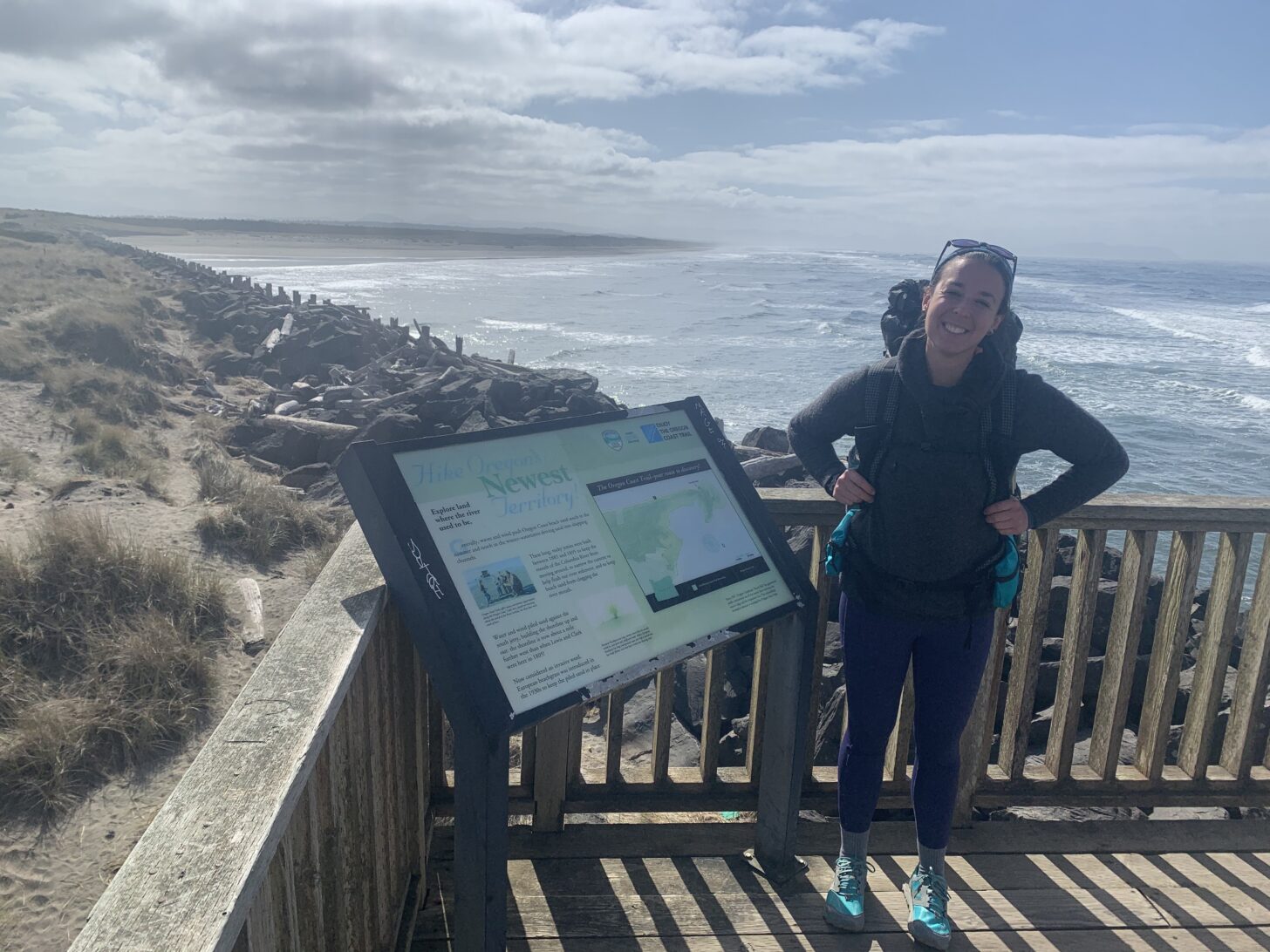 a woman stands next to a trail sign for the Oregon Coast Trail with the beach and ocean in the background.