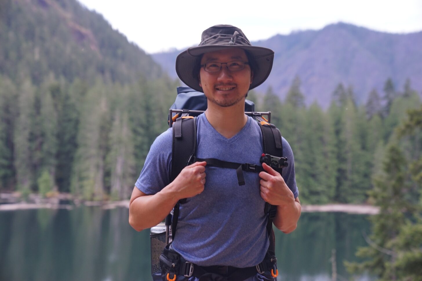 A man stands facing the camera with a lake and trees in the background. He is wearing the Vargo ExoTi BOG backpack.