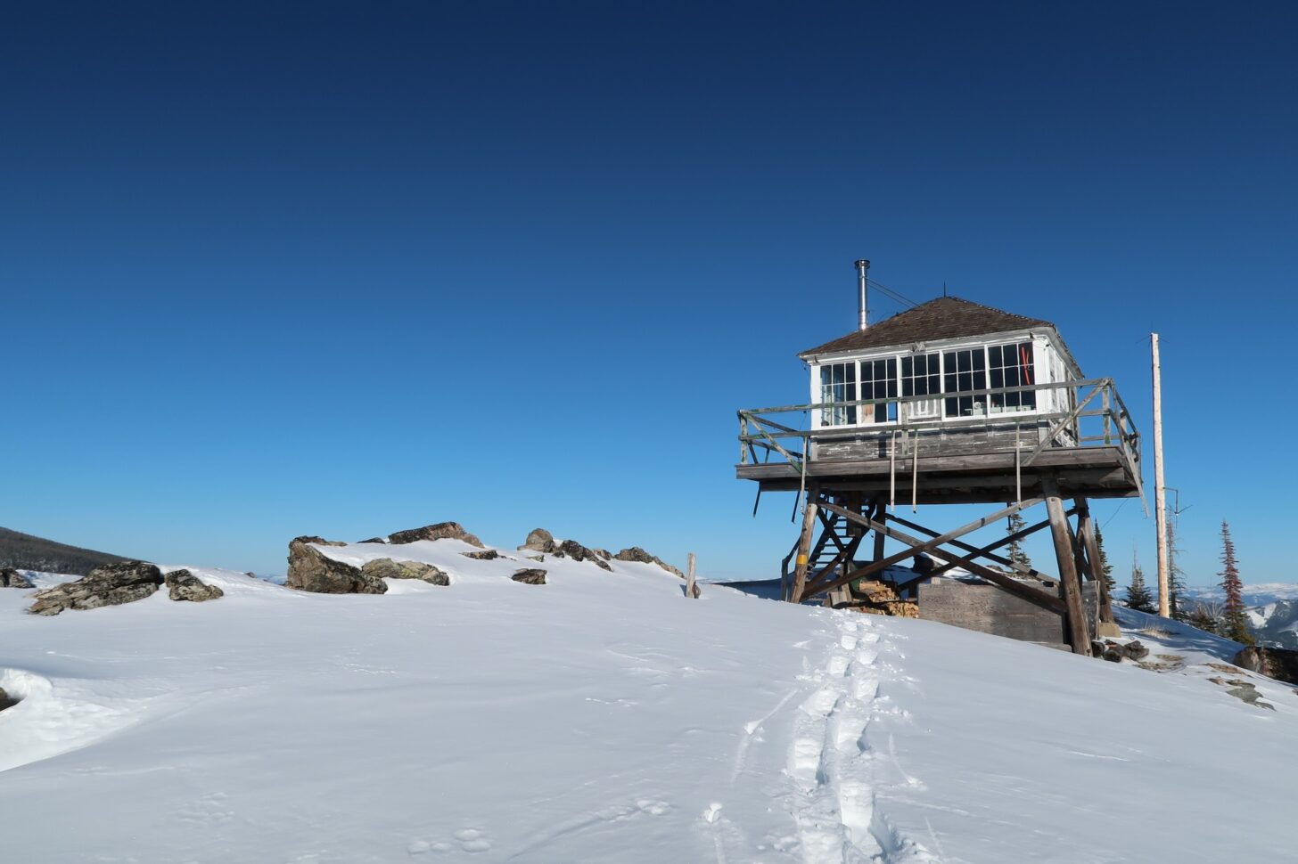 Image shows a fire lookout building on top of a mountain with snowshoe tracks leading up to it.