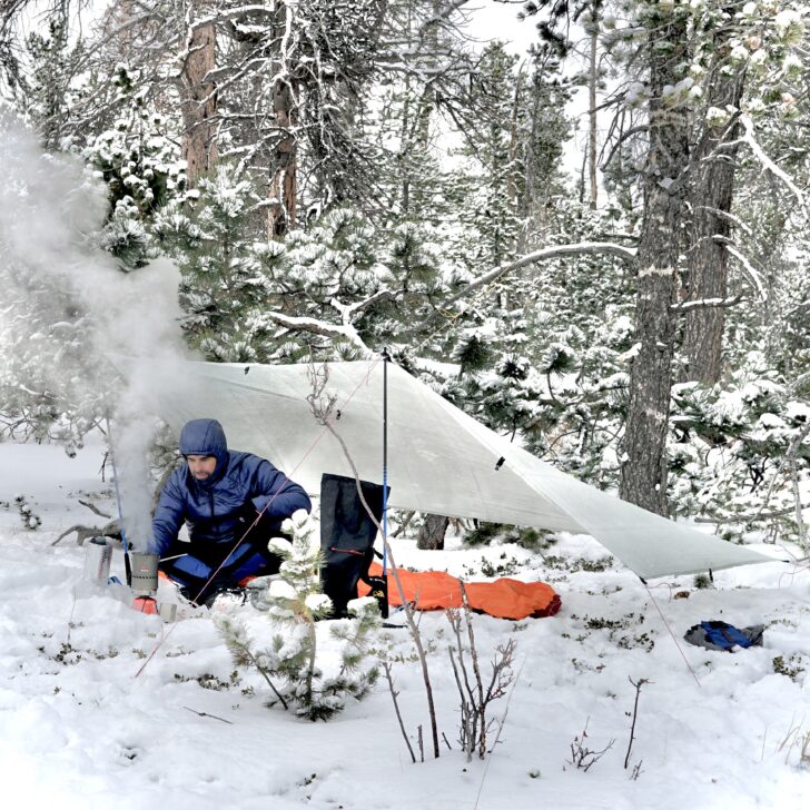 hiker operating a stove while camping under a tarp in a snowy forest