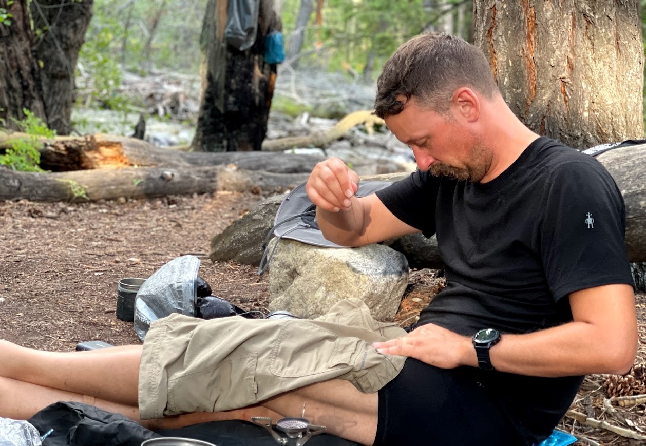 a man sits on the ground using a needle and thread to repair a pair of shorts