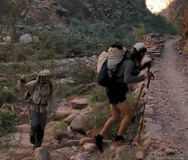 two men wearing packs climb up onto a trail from a rocky area
