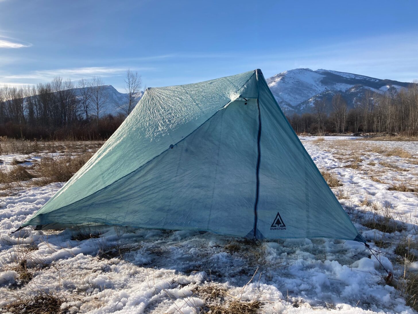 A tent and background are pitched on top of snow with mountains in the background.