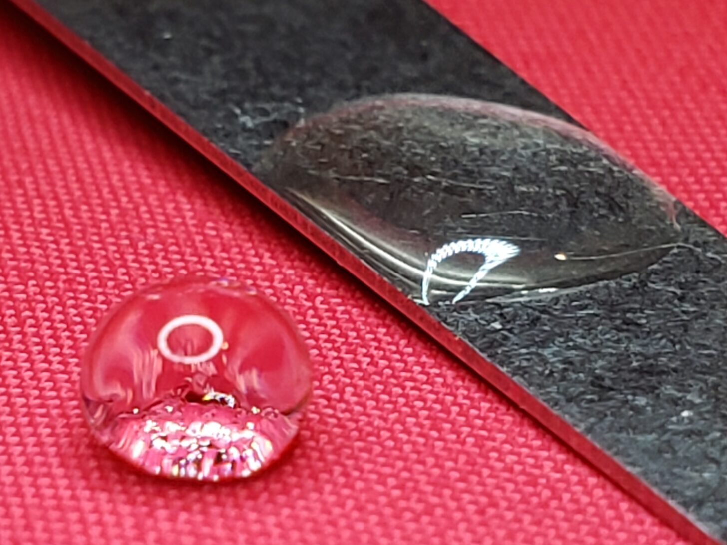 The red fabric is a Neoshell fabric. It is treated with DWR to render its surface highly hydrophobic. As a result, the left drop of water is nearly spherical. The contact angle, viewed at the bottom of the drop, is well over 90°. The flattening at the bottom is due either to the force of gravity or a low level of adhesive force. One way to determine this is to invert the surface. If the drop remains attached, there is some adhesive force. If the drop slides off, the flattening is likely due to gravity. The right drop is visibly flat. The contact angle is very low, perhaps 20°. We can see that this surface is hydrophilic. The water molecules show a high level of adhesion to the metal surface. In fact, I flipped the metal strip upside down and the drop remained in place! If you think metal is necessarily hydrophobic, think again.