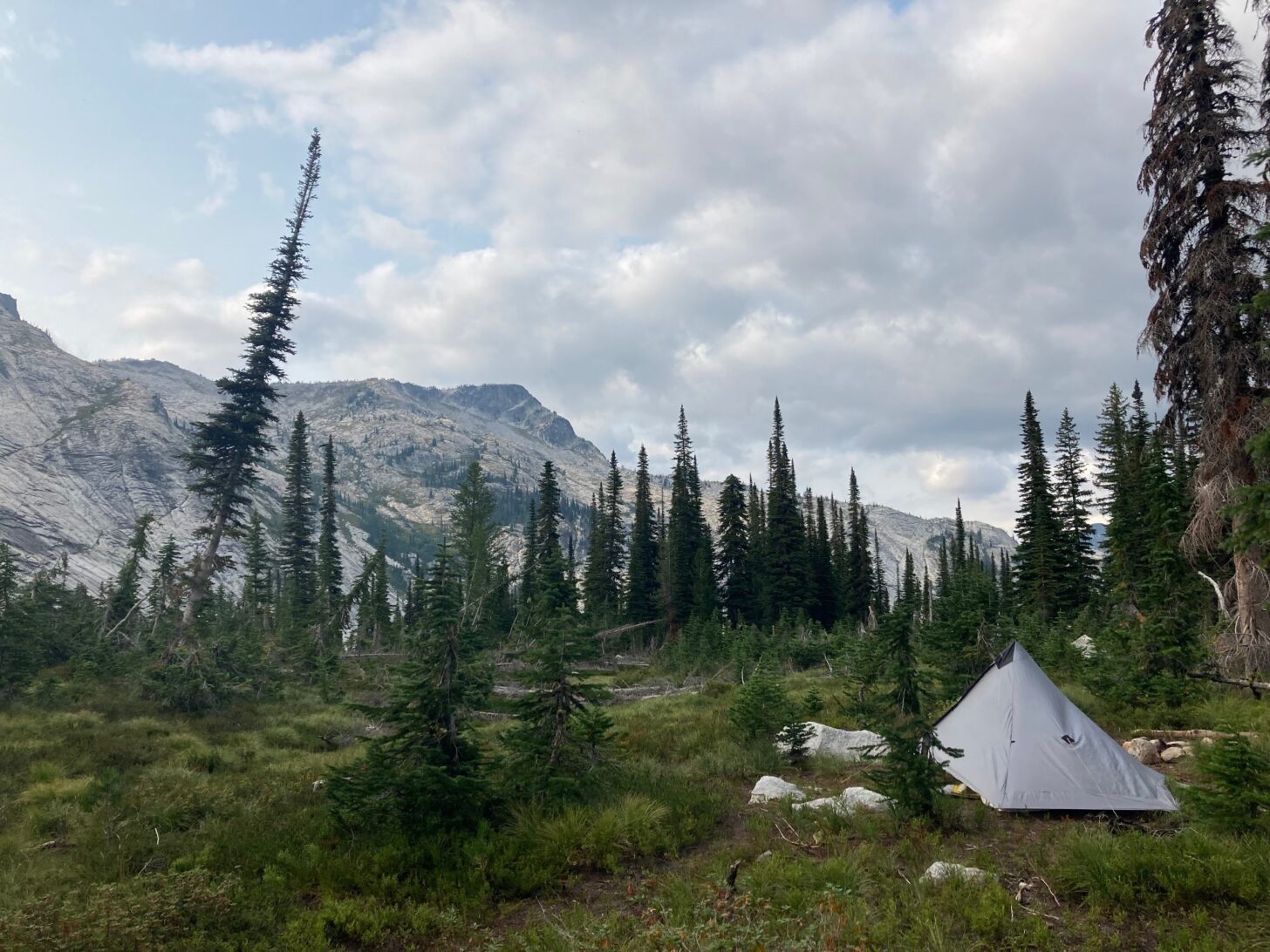 A tent in a subalpine forest in the foreground, with mountains in the background and clouds above.