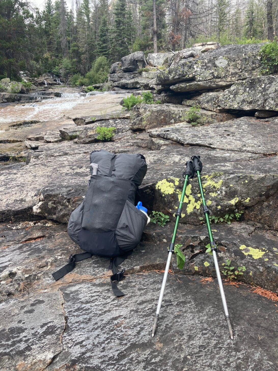 A backpack and trekking poles are centered in this photo with a stream in the background.