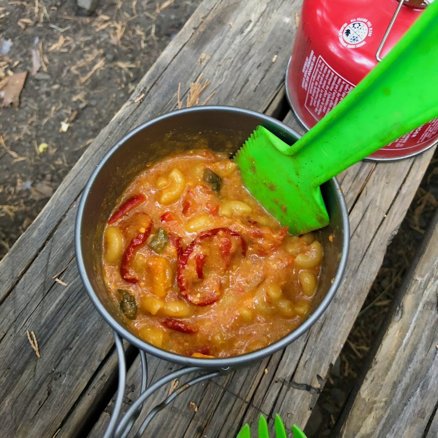 a shot from above, looking down into a pot of food. The food is a hearty-looking tomato sauce with macaroni noodles and cheese. A green plastic utensil sticks out of the pot.