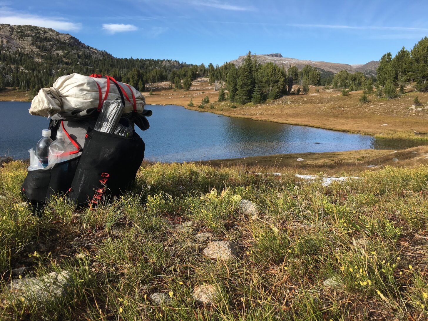 a backpack sits on a hill above an alpine lake. Two water bottles are crammed in the side pocket.