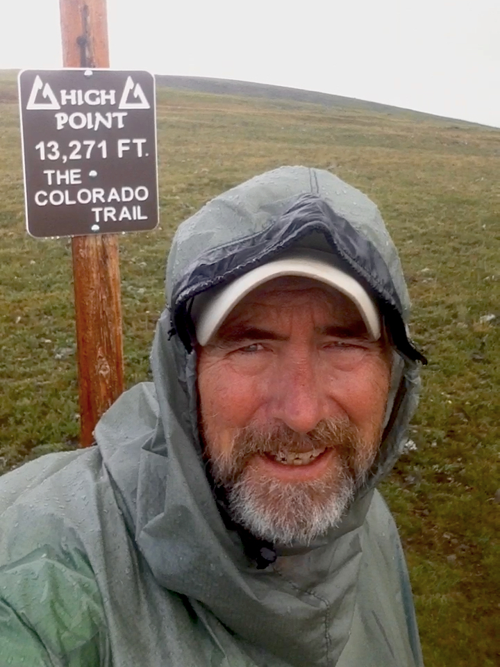 A man wearing a poncho takes a selfie next to a sign. The sign text reads "High point, 13,271 feet, the Colorado Trail."