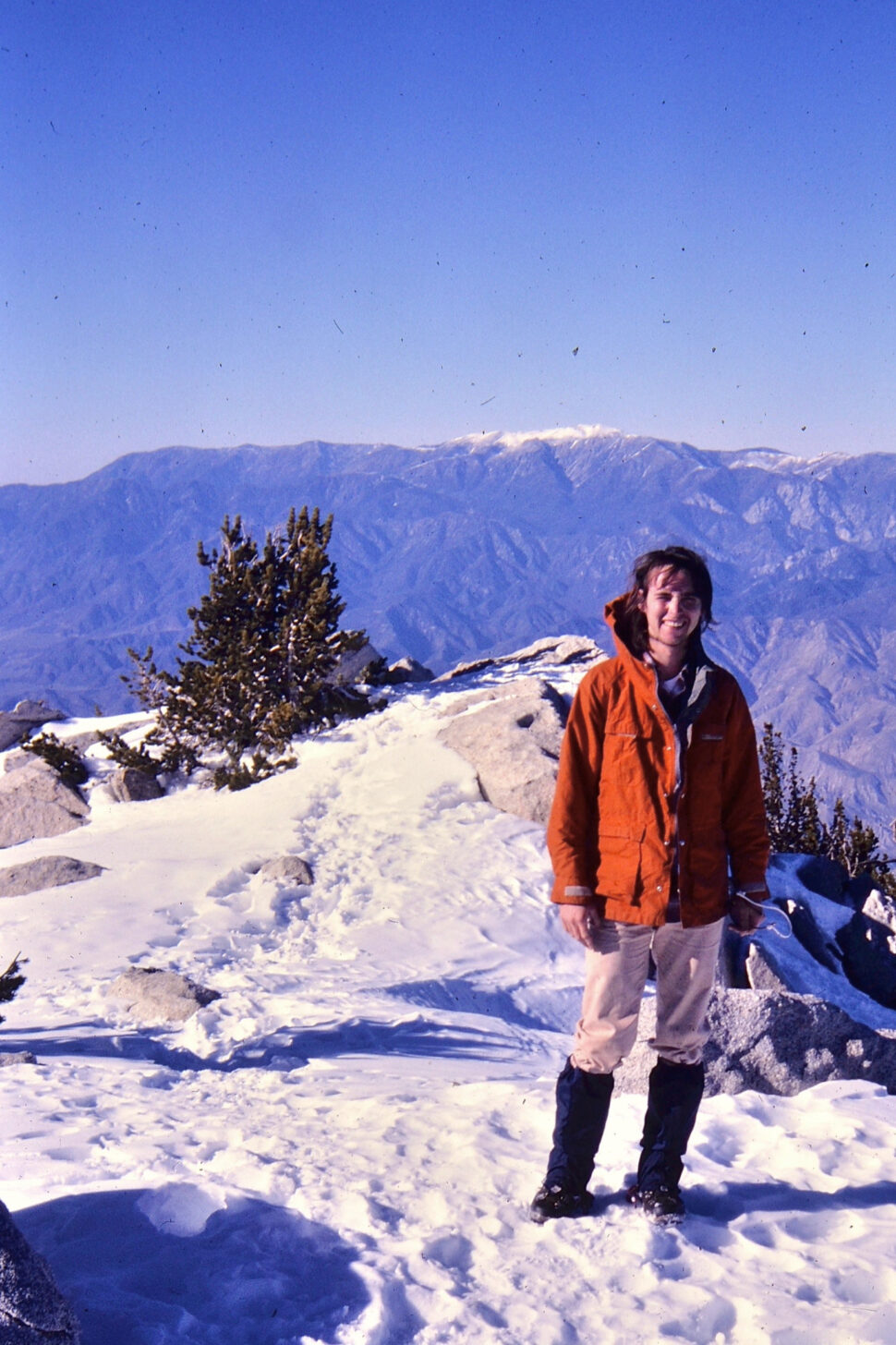 A young man stands on a snowy peak with mountains in the background while wearing an orange jacket.