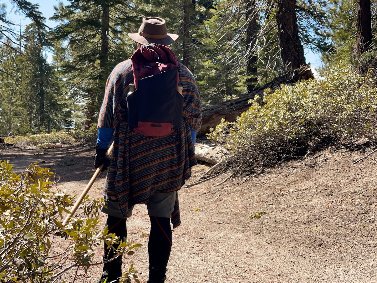 A man wearing the Atom Packs Nanu in addition to a great hat walks away from the camera along a forested road.