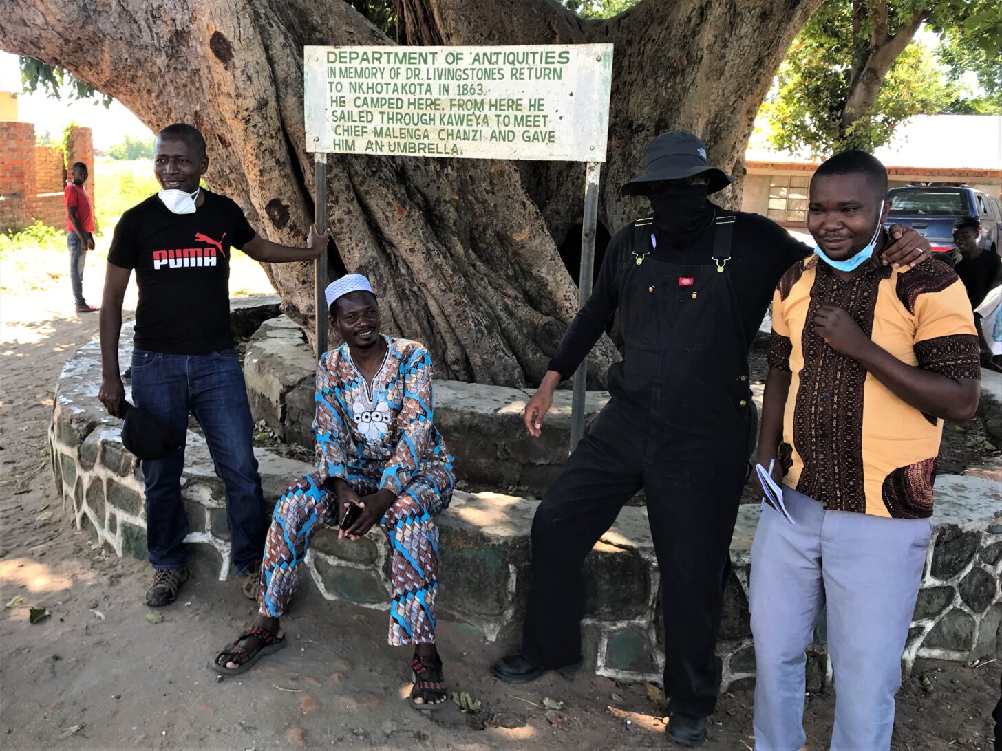 A group of people stand next to a sign. Sign reads Department of Antiquities: In memory of Dr. Livingston&squot;s return to Nkhotakota in 1863. He camped here. From here he sailed through Kaweya to meet Chief Malenga Chanzi and gave him an umbrella."
