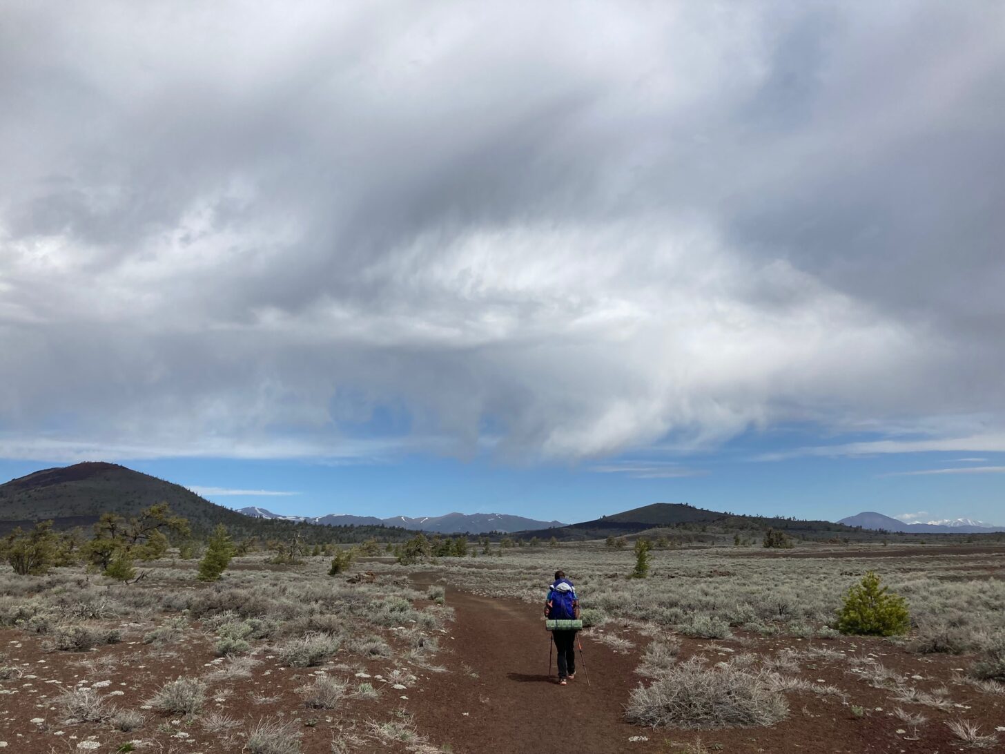 A backpacker hikes through a sagebrush flat with mountains in the distance and clouds above.