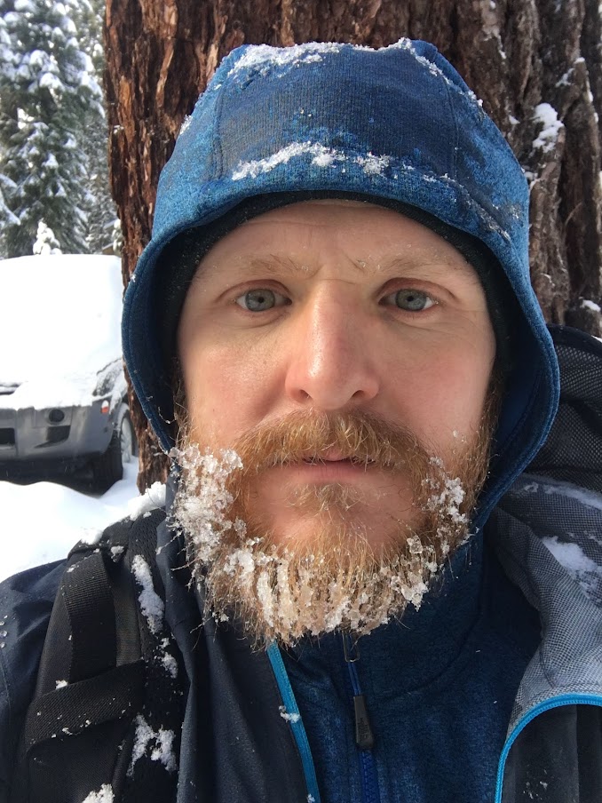A man with a short, unimpressive beard covered in ice looks at the camera. In the background, a snow-covered car.