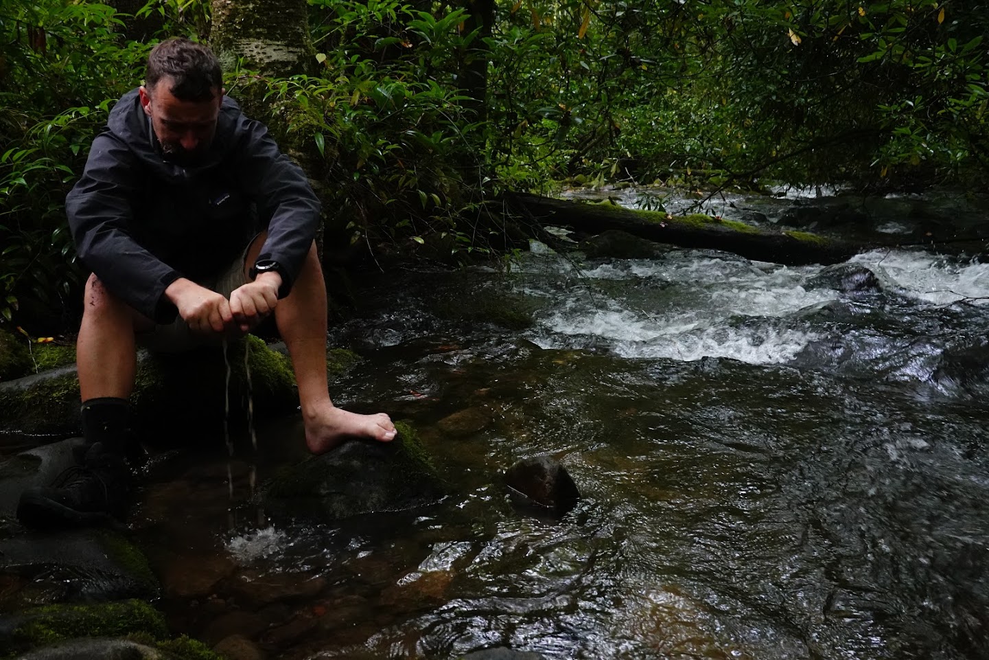 a man squeezes out a wet sock while sitting on a rock next to a creek.