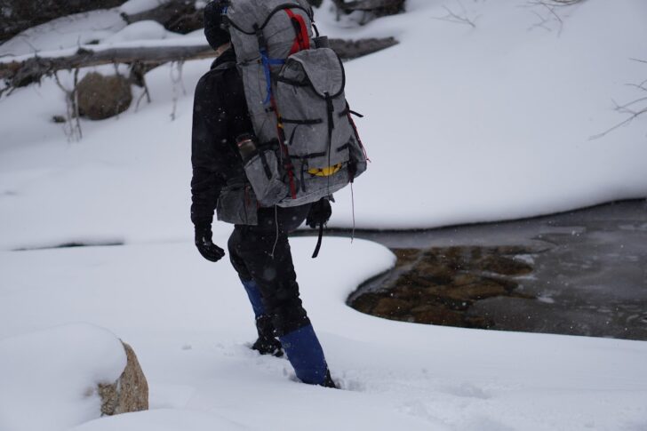 a man walks through deep snow near a frozen creek while wearing a backpack adorned with lots of straps and buckles.