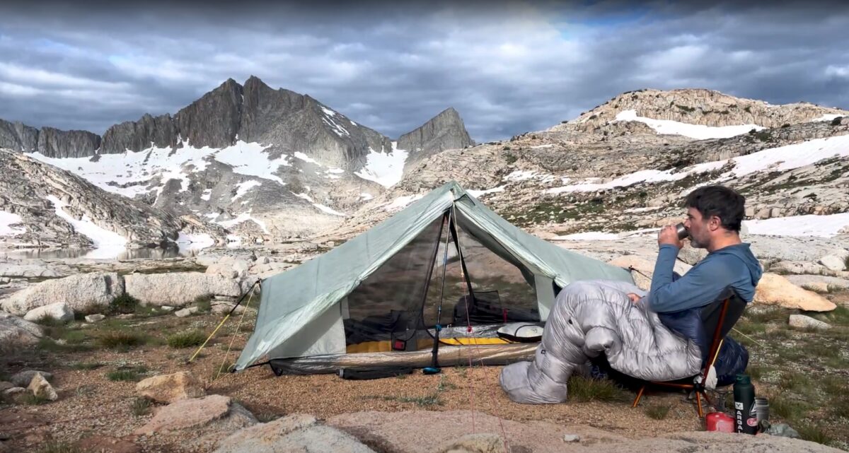 man drinking coffee by a tent in the mountains