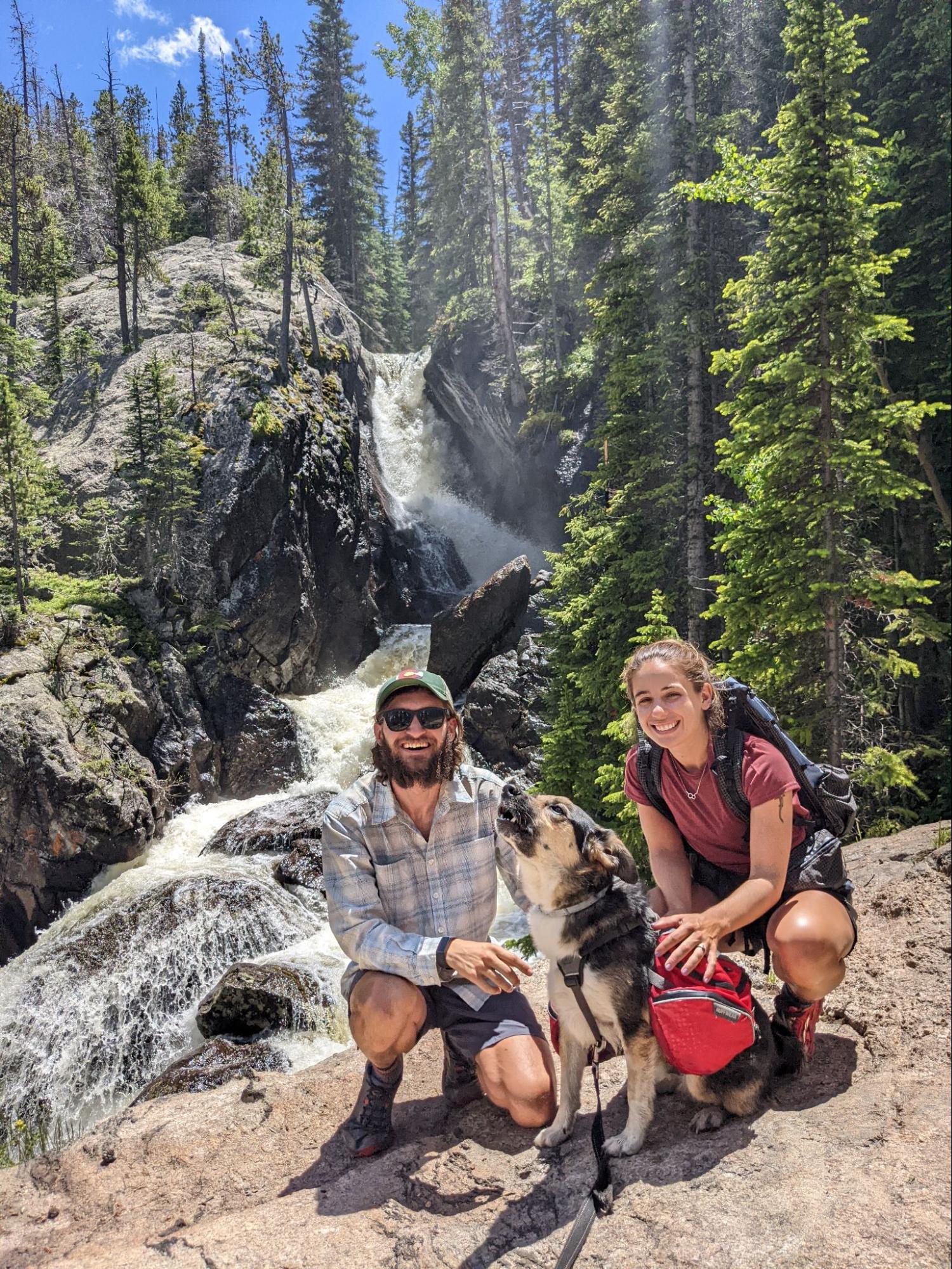 a man and a woman pose with a dog in front of a waterfall