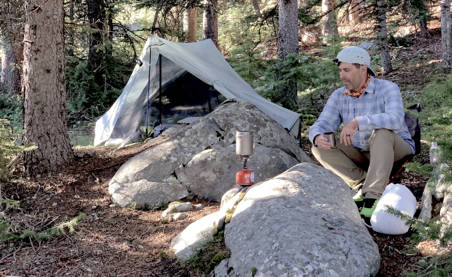 man sitting by a tent drinking tea in the woods